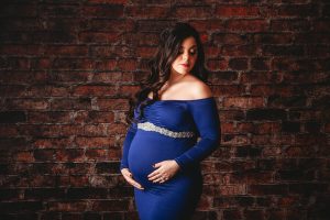 Pregnant woman in blue dress posing against brick wall.