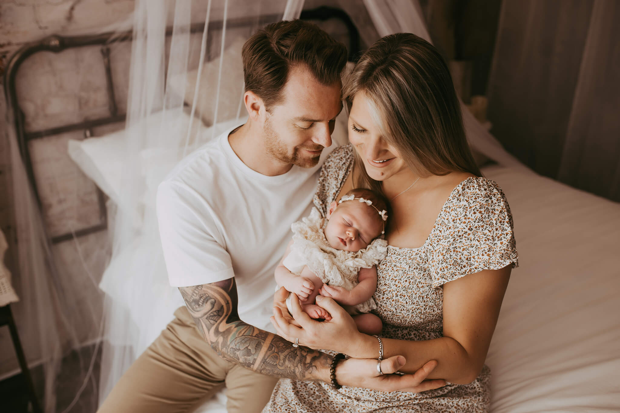 New parents sit on a bed hugging while mom holds their newborn baby girl in her arms