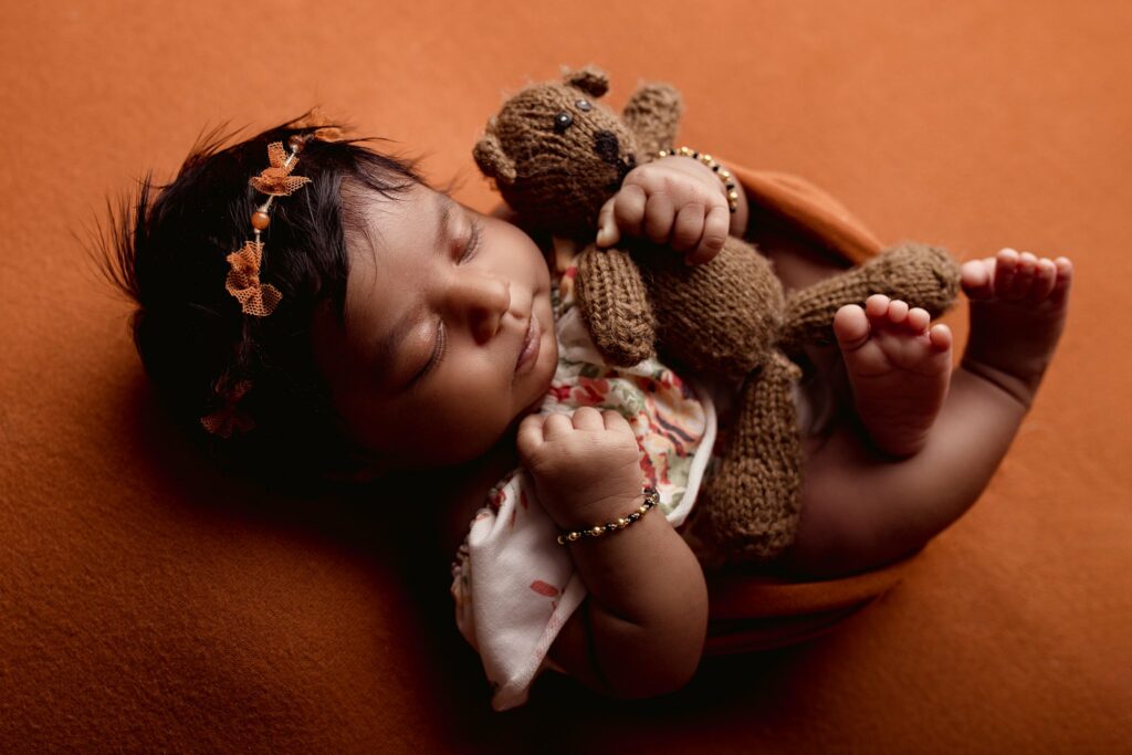 A newborn baby sleeps in an orange swaddle cuddling a knit teddy bear