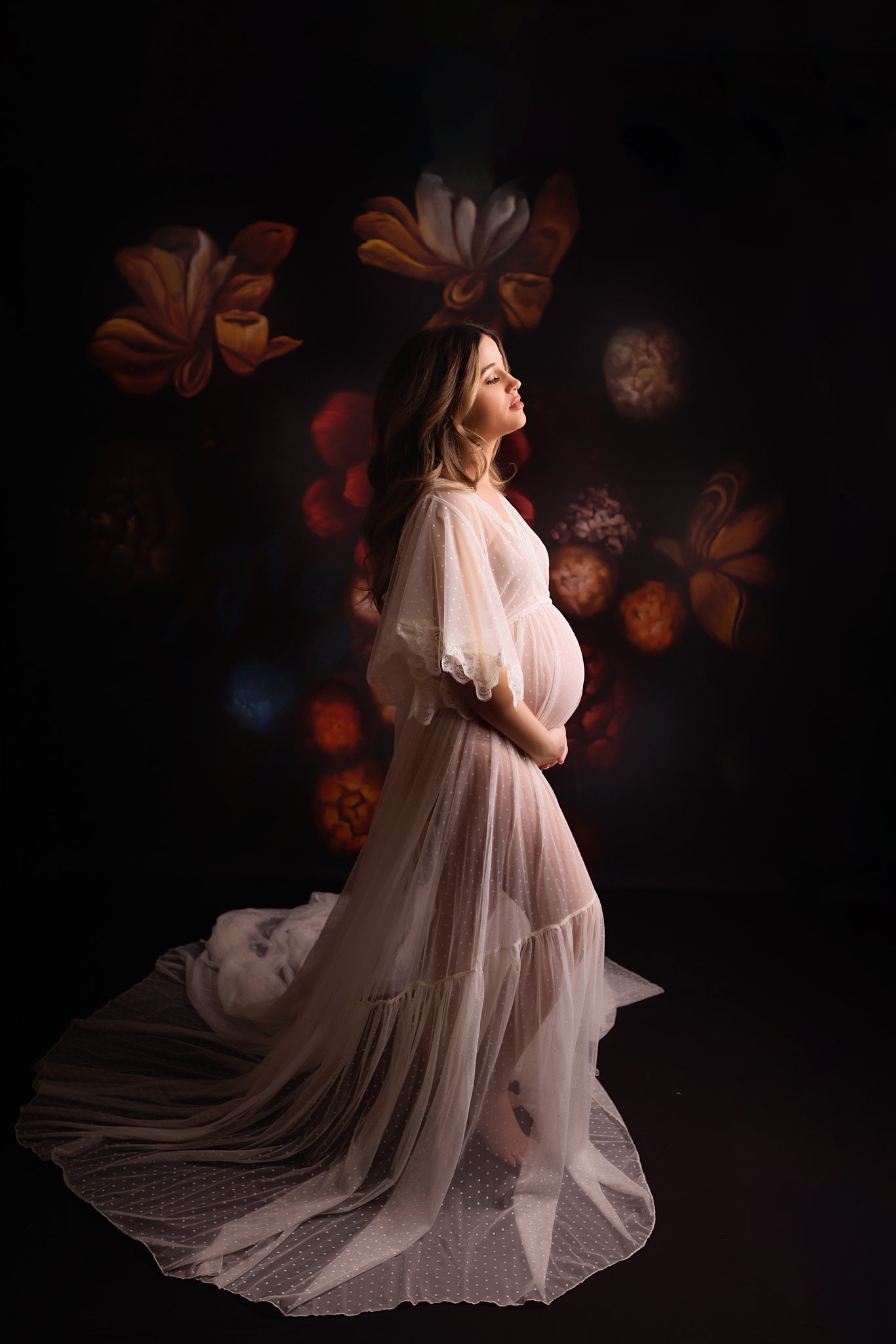 Mother to be stands in a studio with a dark floral backdrop in a sheer maternity gown prenatal yoga toronto