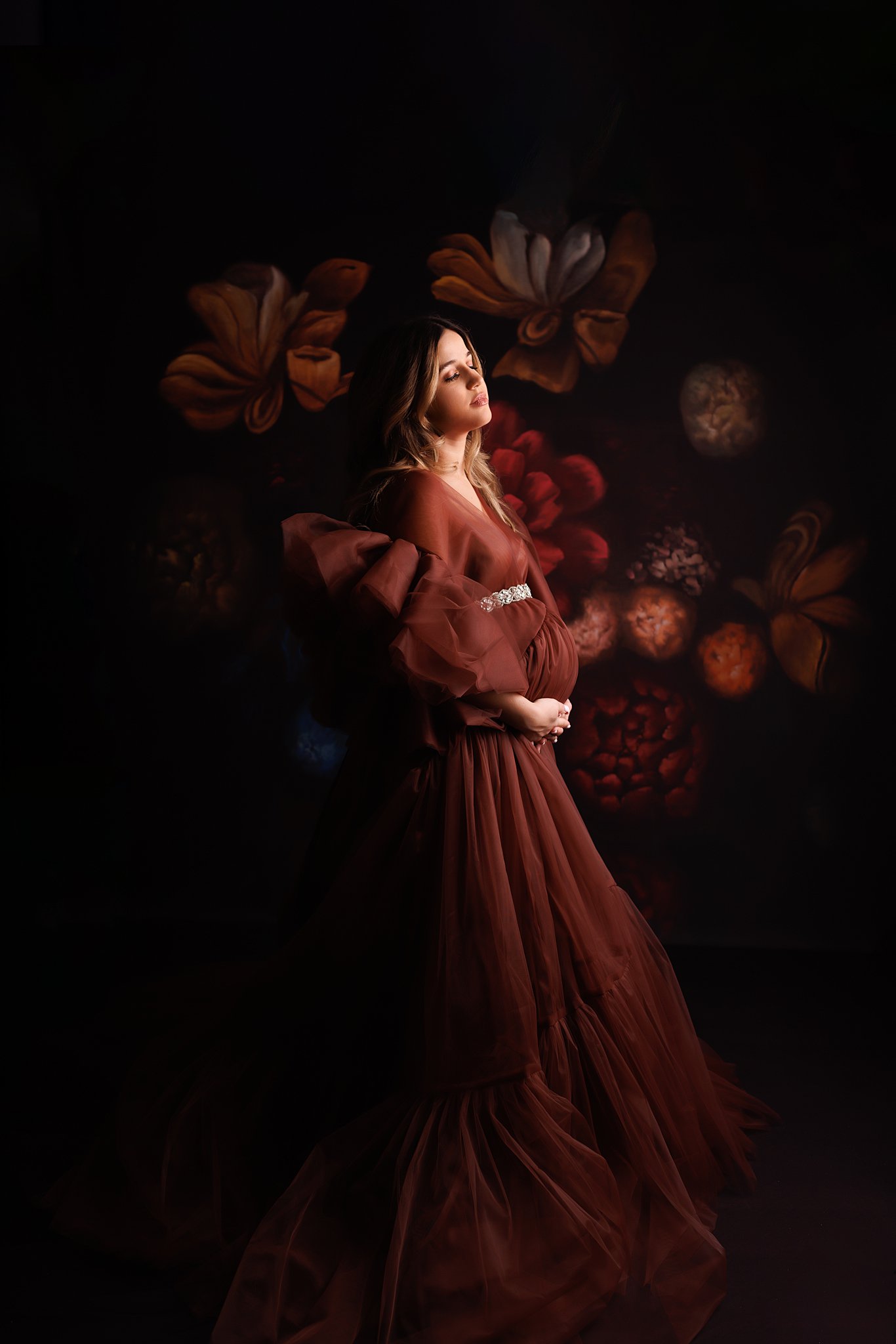 A mother to be stands in a studio wearing a maroon dress with a dark floral background serenity birth studio barrie