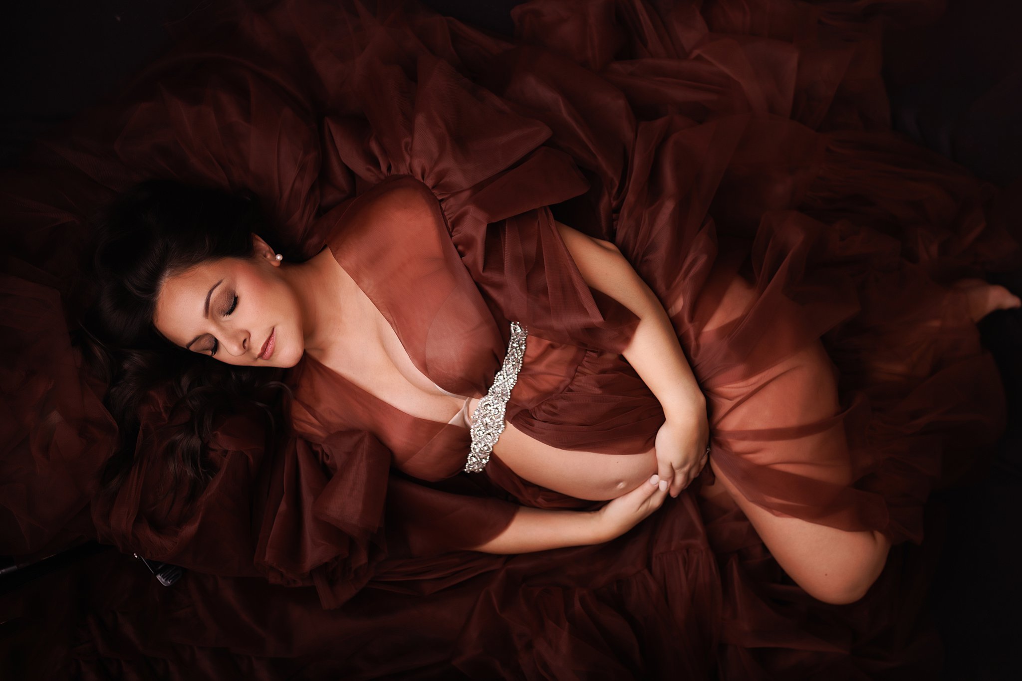 A mother to be in a large flowing burgundy maternity gown lays on floor holding her bump with a diamond belt toronto prenatal classes