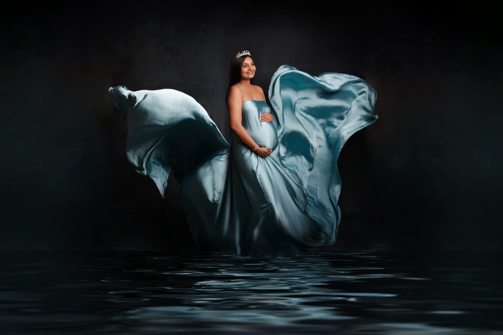 A mother to be wearing a blue silk maternity gown that is flowing around her